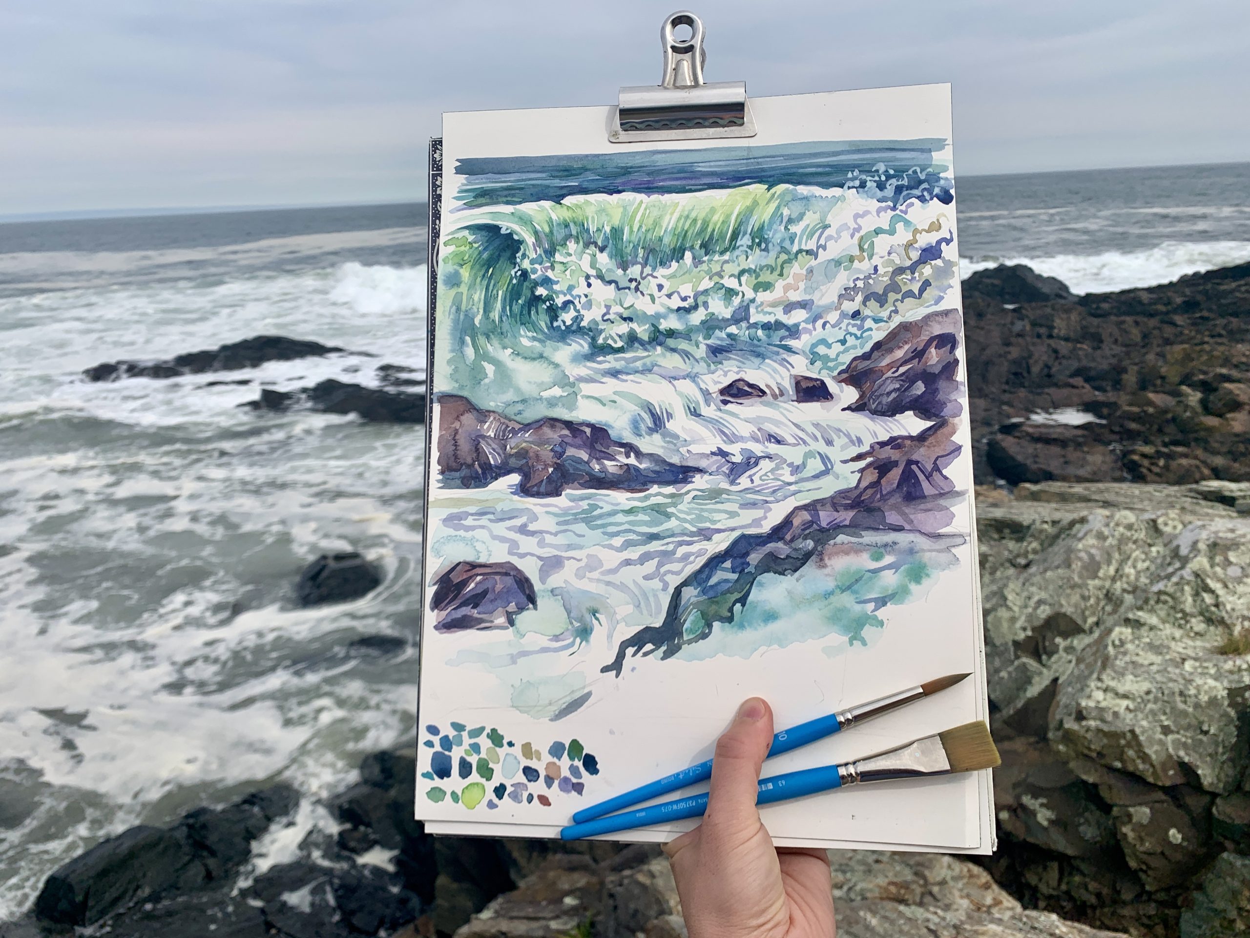 Photograph of the ocean with a hand holding a watercolor picture of the water and paintbrushes